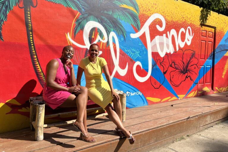 Ivana and Adalia, both Black women, sit on a bench in front of a wall with a colorful mural that reads "El Tunco"