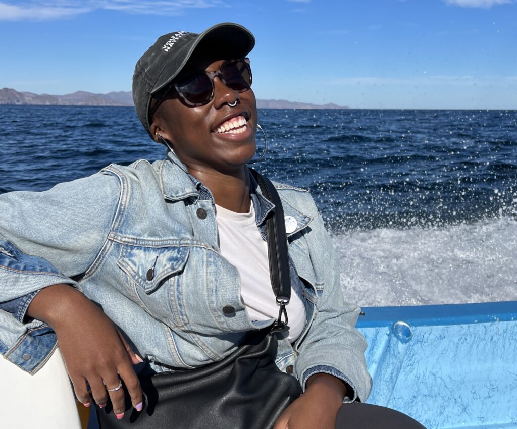 Ivana, a Black woman, is wearing a black cap, sunglasses, and light-wash jean jacket. She's sitting on a boat with one arm back, resting on the side and water spalshing behind her. Her head is tilted back and she's smiling a full smile at the camera.