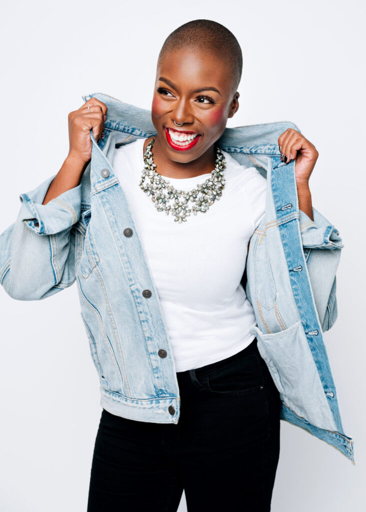 Ivana, a Black woman, is shrugging on a light-wash denim jacket over her white t-shirt. She's smiling wide and looking to the left of the viewer.