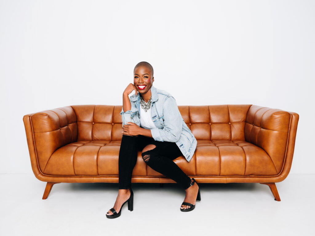 Ivana, a Black woman, is sitting on a brown, tufted leather couch and looking at the camera with a big smile while lightly resting her cheek on her hand.
