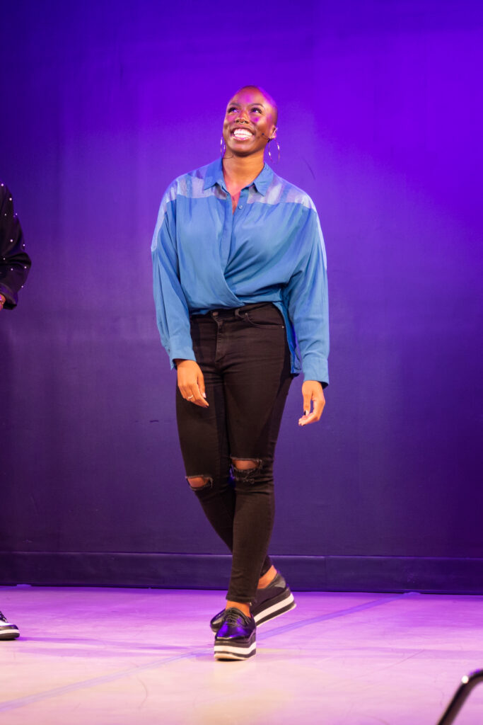 A full body shot of Ivana, a Black woman, walking toward the camera with her head tilted back as if she's mid-laugh. She's wearing a chambray shirt and black pants with tears at the knees.