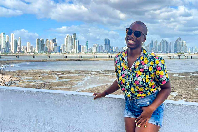Ivana, a Black woman, stands in front of a white half wall with a city skyline in the background. There's a long bridge visible over water and the tide is out revealing some mud flats just beyond the wall. Ivana is smiling and wearing a button up shirt with a pineapple print and jean shorts.