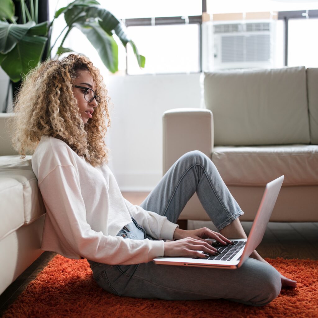 A person with shoulder length curly hair is sitting on the floor, leaning back against a couch. They have a laptop resting on one leg curled in front of them and the other is bent upwards. They are working on the computer.